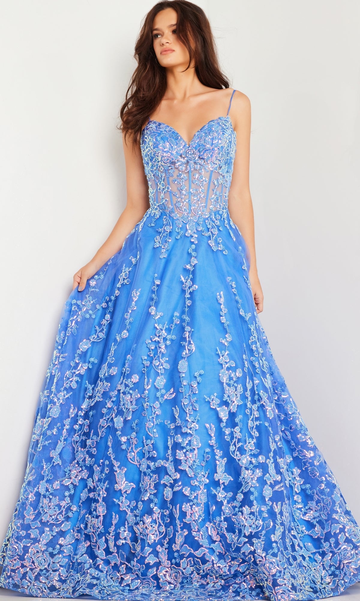 A woman wears the Long Prom Dress 29072 by Jovani, a turquoise blue ball gown with floral sequin embroidery, fitted bodice, thin straps, and a flared skirt, standing against a plain light background.